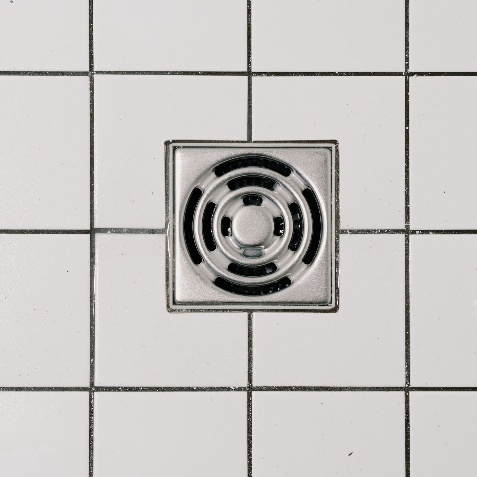 Siphon carré en inox posé exactement dans la trame des joints d’une faïence blanche