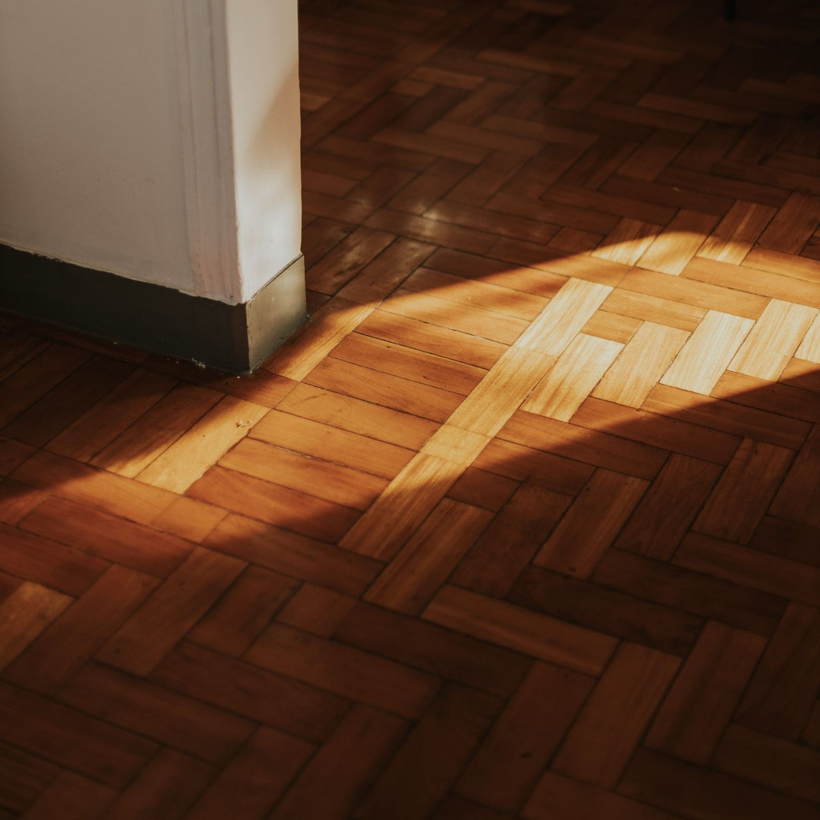 Parquet à bâtons rompus éclairé par une lumière rasante, au pied d’un mur blanc